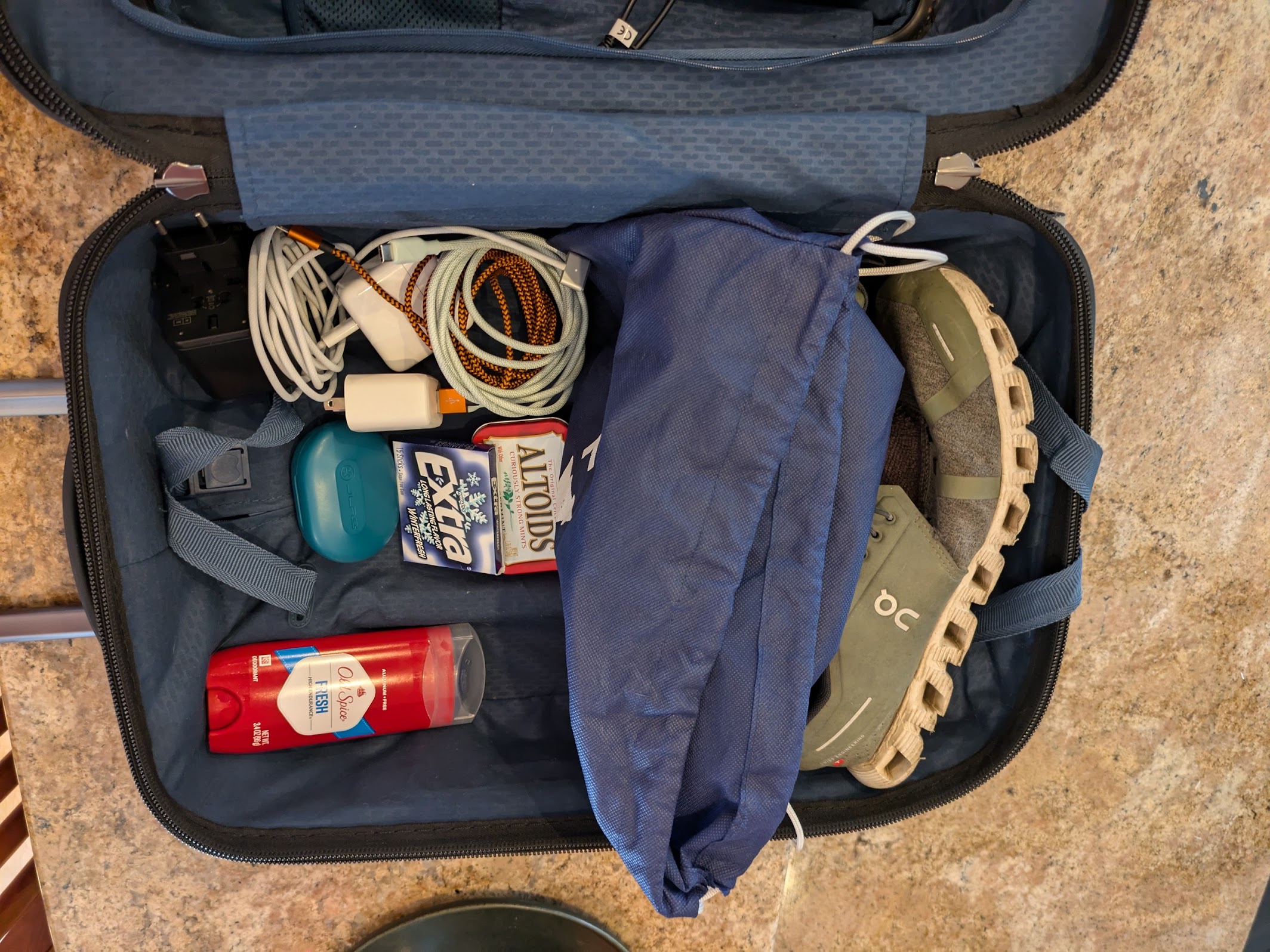 Packed in a small suitcase: On running shoes, THAT Conf blue tote bag, Extra gum, Altoids mints, Jlab wireless headphones case, Old Spice Fresh deodorant, Macbook charger, international power adapter, and USB A and C cables in a wall plug.